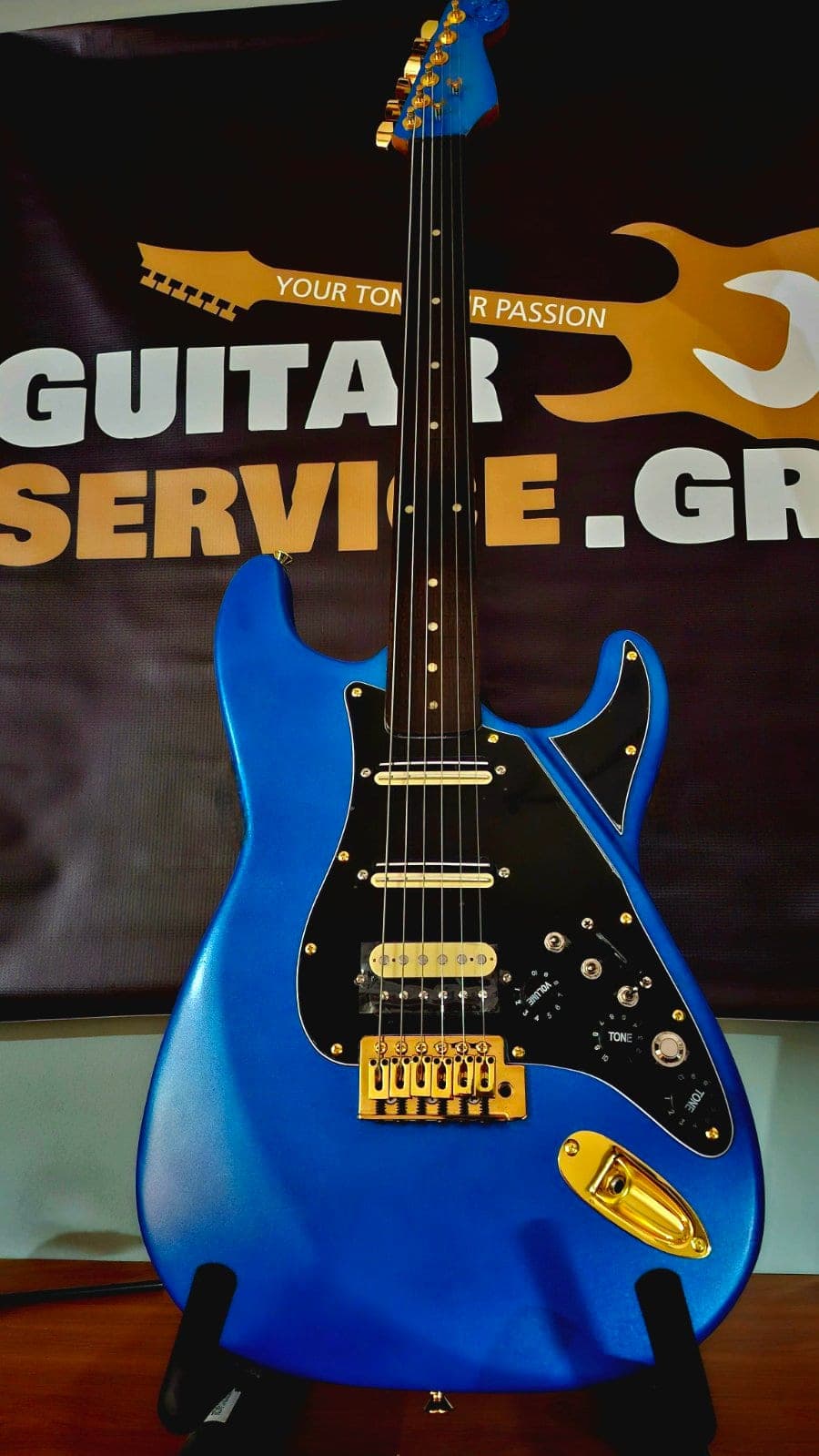 Μπλε ηλεκτρική κιθάρα τύπου Stratocaster με χρυσά εξαρτήματα, σε βάση μπροστά από το banner του εργαστηρίου.
